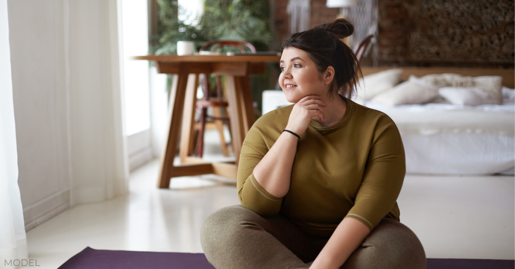 Charming plus size woman in sportswear sitting cross legged on mat, going to practice yoga, smiling (model)