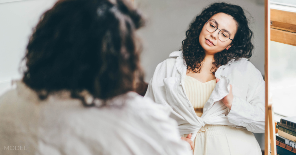 Curly haired overweight young woman in glasses ties white shirt and admires choice standing in front of large mirror in stylish room reflection view (model)