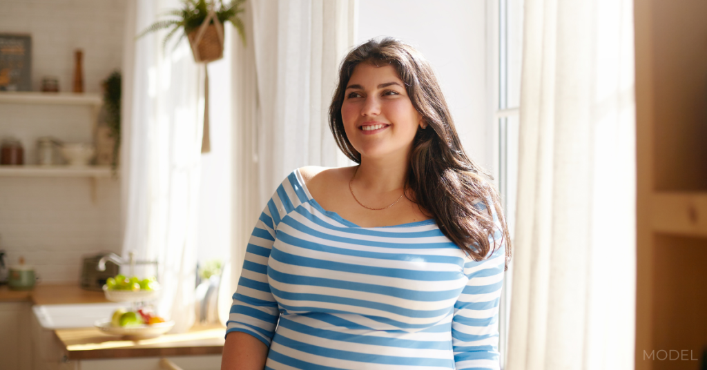 Indoor shot of gorgeous brunette Latin girl posing by window in kitchen dressed in xxl blue and white striped shirt, having joyful beaming smile (model)