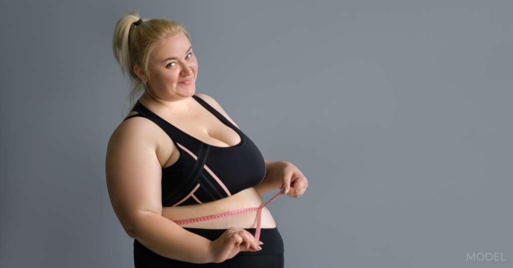 Plus size lady measuring her waist with a measuring tape on a gray studio background (model)