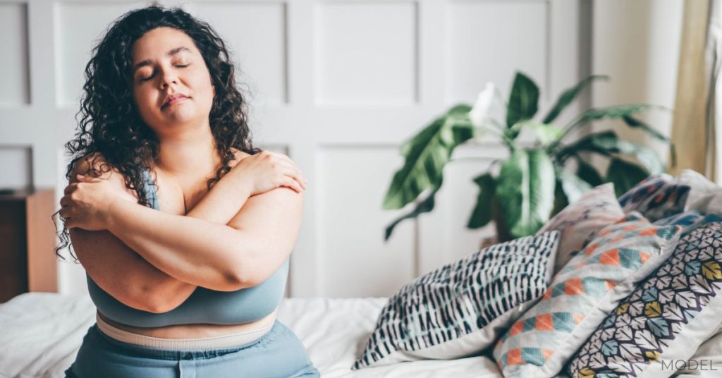 Curly haired overweight young woman in blue top and shorts in stylish bedroom. (Model)