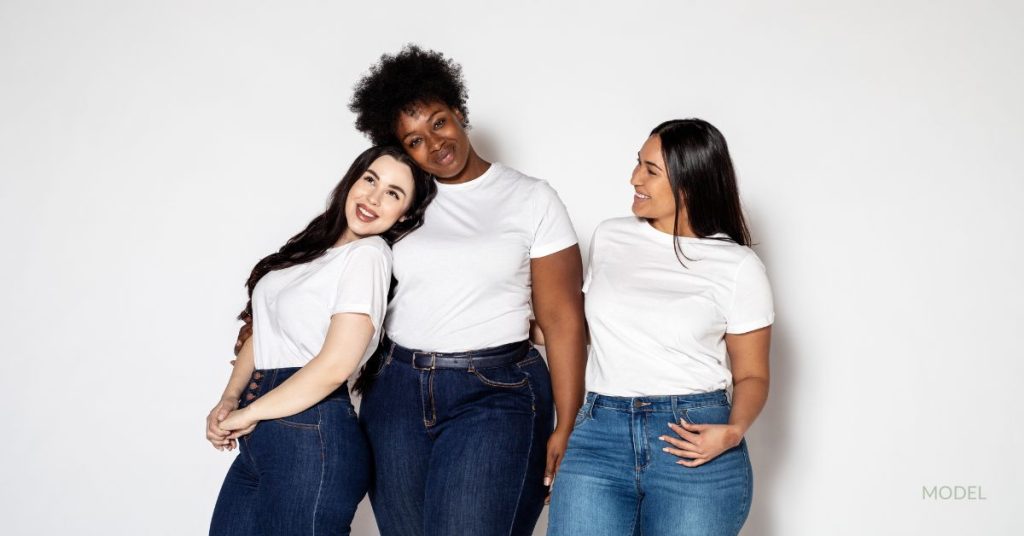 Three plus-sized women in jeans and white t-shirts are embracing each other while smiling at each other and the camera. (Models)