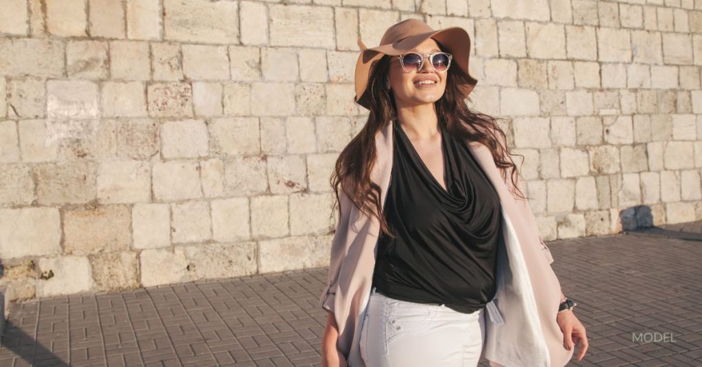A beautiful plus-sized woman in a sun hat and sunglasses is smiling while on a walk outside. (Model)