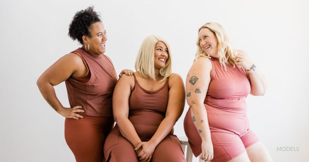 Three plus-sized woman in brown and pink workout clothing are huddled together laughing and smiling. (Models)